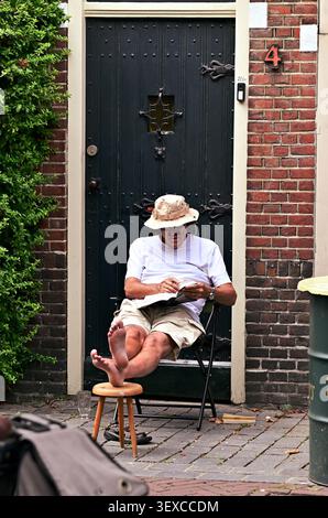 Barfußer Mann liest ein Buch vor einem traditionellen niederländischen Haus, während in der Nähe ein lokales Straßenevent stattfindet Stockfoto