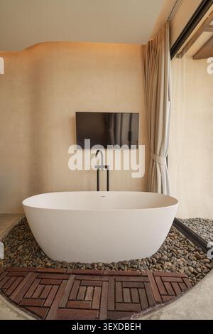 Elegante freistehende weiße Badewanne auf Kieselboden mit Fernseher auf Sandsteinhintergrund in tropischer Villa Stockfoto