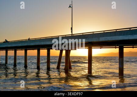 Blick auf den Venice Fishing Pier, der sich bei Sonnenuntergang über dem Pazifischen Ozean erstreckt, mit goldenem Licht, das auf dem Wasser reflektiert, und Silhouetten der Menschen in Mar Stockfoto
