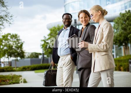 Drei Profis laufen gemeinsam auf einem Stadtpfad unter bewölktem Himmel. Sie führen eine Diskussion, während sie sich ein Tablet ansehen. Die Szene zeigt ein Ur Stockfoto