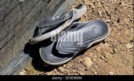 Ein hoher Blickwinkel auf ein Paar alte, schlammgebeizte schwarze Flip-Flop-Sandalen aus Gummi, die auf trockenem, holprigem Schmutz entsorgt wurden, neben einer grauen Netzkonstruktion Stockfoto