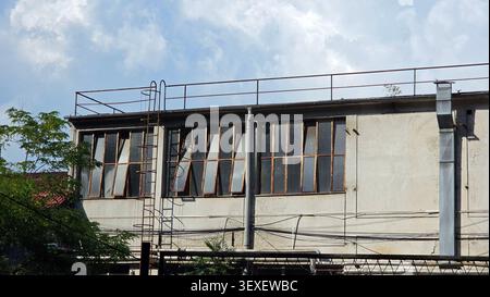 Die Außenfassade einer verwitterten Betonfabrik verfügt über eine Reihe offener Stahlrahmenfenster, eine eiserne Serviceleiter und Dachgeländer unter bewölktem Himmel Stockfoto