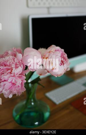Heller kreativer Arbeitsbereich mit frischen Pfingstrosen, Desktop-Computer und Holzschreibtisch, ideal für Produktivität und Fernarbeit. Stockfoto