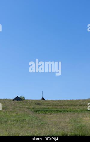 Eine minimalistische ländliche Landschaft mit einer kleinen hölzernen Scheune und einem traditionellen Heuhaufen auf einem grünen Grashügel unter einem weitläufigen blauen Himmel mit reichlich Überfluss Stockfoto