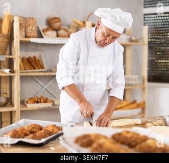 Senior man arbeitet in der Bäckerei als Bäcker, schneidet Teig in Portionen, formt Teigstücke, um Brötchen zu kreieren. Stockfoto