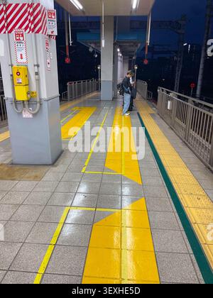Person, die am Abend auf einem Bahnsteig mit gelben Sicherheitsleinen und hellen Bodenmarkierungen an einem japanischen Bahnhof steht. Fukuoka, Japan – November Stockfoto