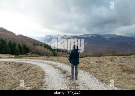 Ein einsamer Reisender in einer Winterjacke steht auf einer Feldstraße auf dem Monte Zurrone und genießt den kalten Tag und den epischen Überblick über den massiven Apennin-berg Stockfoto