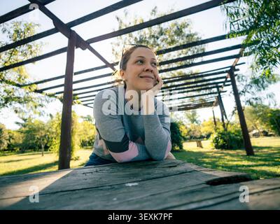 Die junge Frau ruht auf einem Holztisch. Sie lächelt nachdenklich, Hand nahe dem Kinn. Die Pergola aus Metall wirft Schatten auf die Szene. Sonnenlicht filtert durch das Blattwerk Stockfoto
