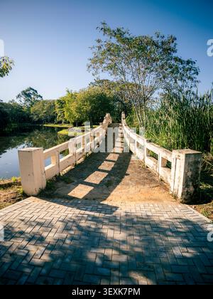 Eine Steinbrücke überspannt einen ruhigen Fluss. Weiße Geländer säumen den Weg. Sonnenlicht wirft Schatten auf den gemauerten Gehweg. Üppige Bäume und Gräser säumen die Szene. Cl Stockfoto