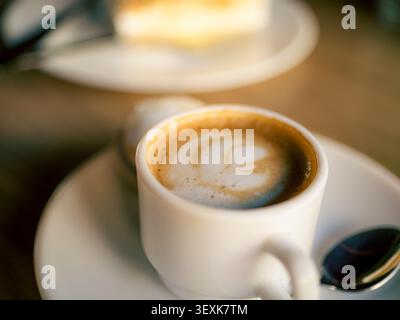 Die weiße Tasse enthält cremige Latte Art. Milchschaum wirbelt im Kaffee. Löffel liegt auf einer Untertasse daneben. Weicher Hintergrund zeigt einen verschwommenen Teig. Diese im Stockfoto