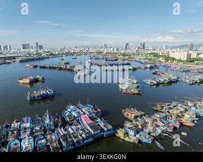 Blick aus der Vogelperspektive auf farbenfrohe Fischerboote im Hafen von da Nang, Vietnam, mit moderner Skyline der Stadt unter klarem blauem Himmel. Stockfoto