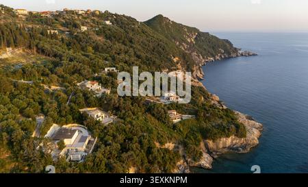 Blick aus der Vogelperspektive auf die Küstenlandschaft mit üppigen grünen Hügeln, verstreuten mediterranen Villen und felsigen Klippen, die auf das tiefblaue Ionische Meer treffen Stockfoto