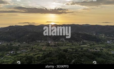 Aus der Vogelperspektive auf die sanften grünen Hügel und die Landschaft von Korfu bei Sonnenuntergang, während die Sonne unter dem Horizont über dem Ionischen Meer in Korfu, Griechenland, eintaucht. Stockfoto