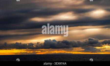 Dramatischer Sonnenuntergangshimmel mit goldenem Licht, das durch dunkle Sturmwolken über dem Ozean bricht Stockfoto
