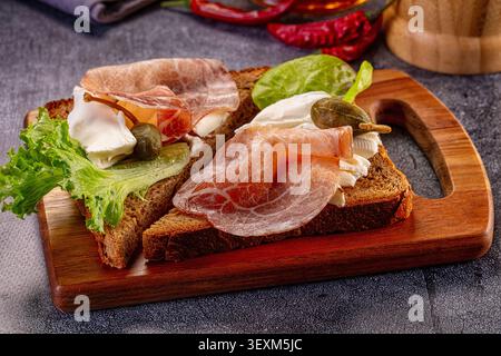 Zwei offene Sandwiches mit Schinken, frischem Mozzarella, knusprigen Salatkapern auf dunklem Brot. Serviert auf rustikalem Holzbrett mit roter Chili-Paprika Stockfoto