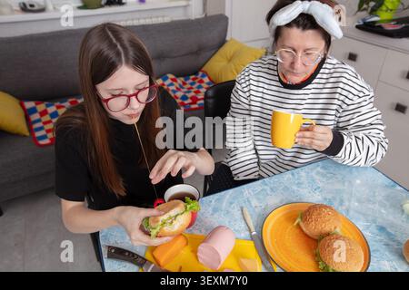 Zwei Freunde sitzen an einem Tisch in einem gemütlichen Wohnzimmer. Einer isst ein Sandwich mit Käse-Stretching, während der andere aus einem gelben Becher schlürft. Stockfoto