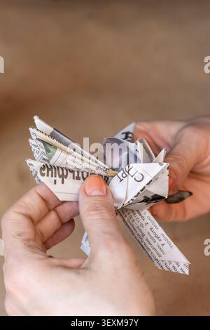 Nahaufnahme einer Person, die aus einer alten Zeitung ein Papierhandwerk herstellt. Die Hände halten das gefaltete Objekt vor einem unscharfen Hintergrund mit Kopierraum. Stockfoto