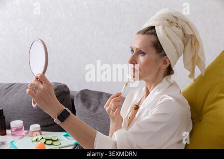 Eine Frau mit einem Handtuch auf dem Kopf untersucht ihre Reflexion in einem Handspiegel, während sie sanft ein Hautpflegewerkzeug auf ihr Gesicht rollt. Sie sitzt in der Komforta Stockfoto