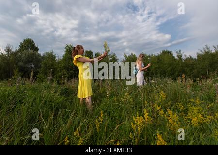 Zwei Frauen in Sommeroutfits lächeln und erkunden ein lebhaftes Wildblumenfeld. Sie interagieren mit der Natur zwischen üppigem Grün unter einer Wolke Stockfoto
