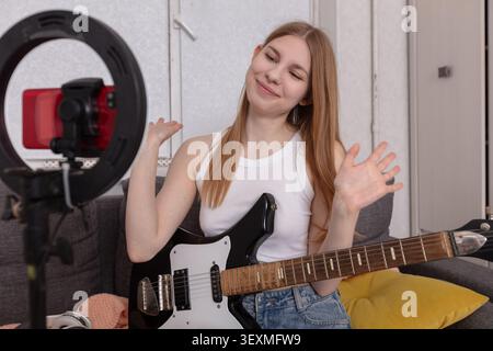 Eine junge Frau mit langen Haaren spielt gern ihre Gitarre, lächelt und interagiert mit den Zuschauern über ihr Smartphone auf einem Stativ Stockfoto