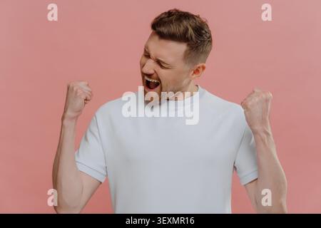 Studio-Porträt eines glücklichen überglückten Mannes in weißem leerem T-Shirt, das Sieg oder Erfolg genießt, aufgeregter Kerl, der Fäuste zusammendrückt und Ja-Geste macht, während er isoliert über pastellrosa Hintergrund steht Stockfoto