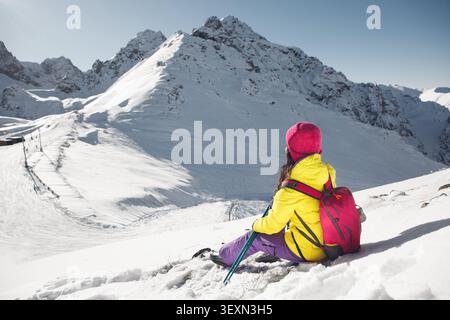 Frau in heller Winterkleidung sitzt auf Snowy Slope und bewundert majestic Mountain View am sonnigen Tag Stockfoto