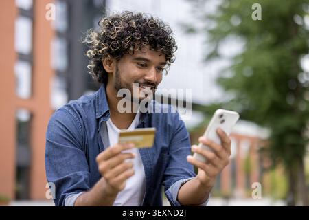 Glücklicher junger Mann mit lockigen Haaren, der sich auf sein Smartphone konzentriert und eine Kreditkarte hält, um im Freien online zu bezahlen oder eine Transaktion zu tätigen Stockfoto