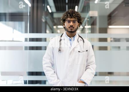Junger indischer Arzt mit lockigen Haaren und Brille, der selbstbewusst in einem modernen Krankenhaus oder Klinikumfeld steht und medizinisches Fachpersonal vertritt Stockfoto