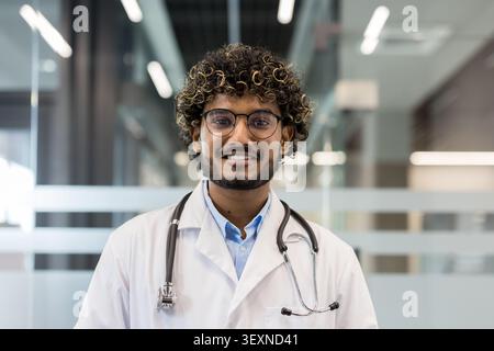 Junger indischer Arzt mit lockigen Haaren und Brille. Mit weißem Labormantel und Stethoskop. Selbstbewusst lächelnd im Krankenhaus oder in der Klinik. Vertretung von professionellen Gesundheitsdiensten und medizinischen Dienstleistungen Stockfoto