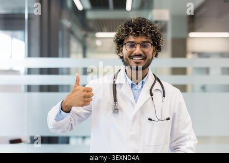Junger männlicher Arzt mit Brille und weißem Labormantel lächelnd und mit Daumen nach oben Geste, symbolisiert Erfolg, Zustimmung, Gesundheit und ausgezeichnete medizinische Versorgung Stockfoto