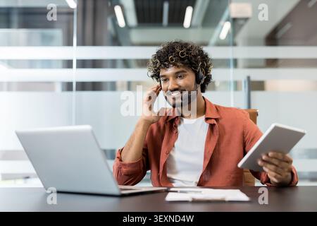 Junger Mann, der als Kundendienstmitarbeiter arbeitet, ein Headset trägt und lächelt, während er ein Notebook und Tablet benutzt, und hilft bei der modernen Büroeinrichtung Stockfoto