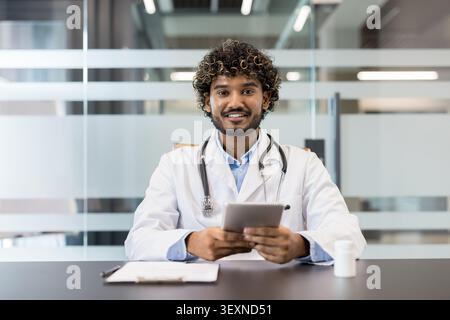 Junger männlicher Arzt mit lockigen Haaren und einem Stethoskop, der einen weißen Labormantel trägt, an einem Schreibtisch sitzt und ein digitales Tablet hält, das moderne Kommunikation und Technologie im Gesundheitswesen repräsentiert Stockfoto