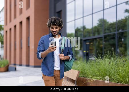 Junger Mann mit einem Rucksack lächelt, während er selbstbewusst geht und sein Handy vor einem modernen Gebäude überprüft, um sich mit der Technologie und der städtischen Umgebung zu verbinden Stockfoto