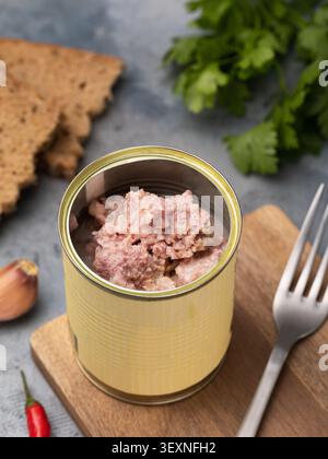 Offene Dose mit Fleischeintopf aus der Dose auf einem Holzbrett, mit Roggenbrot, frischer Petersilie, Zwiebeln und einer Gabel auf einem grauen Tisch Stockfoto