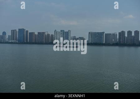 Weitwinkelblick auf einen Frachtkahn, der auf einem breiten Fluss segelt, mit einer modernen Skyline der Stadt und Hochhäusern im Hintergrund. Stockfoto