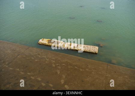 Hochwinkelblick auf einen Betondamm mit einer untergetauchten rechteckigen Steinstruktur im ruhigen, grünen Flusswasser. Stockfoto