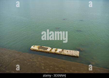 Hochwinkelblick auf einen Betondamm mit einer untergetauchten rechteckigen Steinstruktur im ruhigen, grünen Flusswasser. Stockfoto