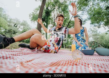 Überraschter junger Mann, der auf eine Picknickdecke fällt, während ein Paar im Sommer ein Picknick im Park genießt Stockfoto