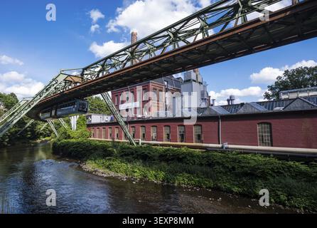 Die Wuppertaler Schwebebahn führt über die Wupper an einem Industriegebäude aus rotem Backstein in Wuppertal vorbei Stockfoto
