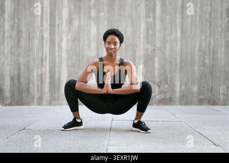 Lächelnde Schwarze Frau mit kurzen schwarzen Haaren, die Sport-BH, Leggings und Sneakers trägt und sich vor einem Workout im Freien auf einem gepflasterten städtischen Bereich vor einer Betonwand ausdehnt. Stockfoto
