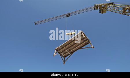 Turmkran Zum Anheben Von Hölzernen Schalungsplattformen. Der Turmkran hebt eine hölzerne Schalungsplattform an, die an Kabeln gegen einen klaren blauen Himmel aufgehängt wird. Stockfoto