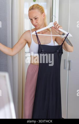 Eine junge Frau, die ein schwarzes Kleid an einem Kleiderbügel hält, vor einem offenen Kleiderschrank steht, über ihre Outfit-Wahl für den Tag nachdenkt und sich darauf vorbereitet Stockfoto