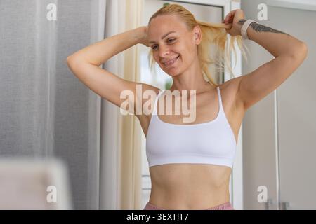 Die blonde Frau stylt ihr Haar zu einem Pferdeschwanz und lächelt selbstbewusst, während sie einen weißen Sport-BH trägt, bereit für ein belebendes Workout und Pre Stockfoto