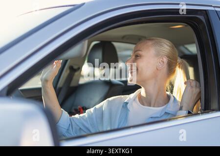Die blonde Fahrerin lächelt, blickt in den Rückspiegel des Autos, passt ihn mit der Hand an und genießt eine fröhliche und komfortable Fahrt Stockfoto