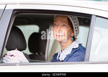 Eine ältere Frau sitzt auf dem Vordersitz eines geparkten Autos und trägt ein traditionelles Kleid mit Spitzendetails und Handschuhen. Sie wirkt nachdenklich, wie sie aussieht Stockfoto