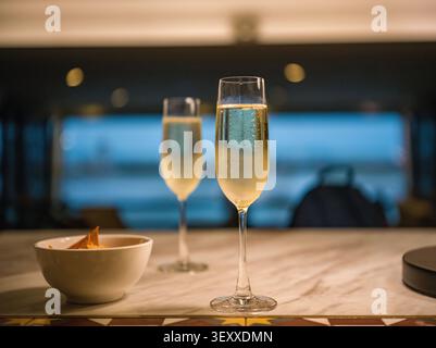 Zwei gefüllte Sektflöten und eine kleine Schüssel mit Snacks auf einer Marmortheke in einer Lounge in der Abenddämmerung. Stockfoto