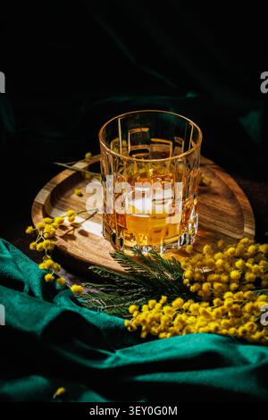 Kristall-Whiskeyglas mit Eis auf Holztablett mit Mimosenblüten für ein Frühlingsgetränk Stockfoto