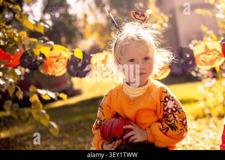 Zwei Kinder genießen Halloween in einem Garten voller Dekoration. Ein Kind hält einen kleinen Kürbis, während es festliche Kleidung trägt. Die Sonne scheint Stockfoto