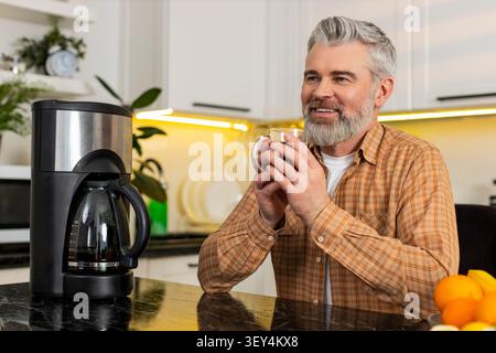 Mann mittleren Alters schlürft frischen Kaffee aus einem klaren Glas Becher in der Küche und fühlt sich glücklich zufrieden und ruhig an einem ruhigen Morgen. Reifer Mann genießt warme Drinks drinnen, reflektiert über Lebensziele Wellness zu Hause. Stockfoto