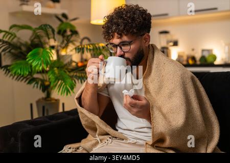 Arabian Mann zu Hause fühlt Halsschmerzen hält Tasse heißen Tee mit Medizin nimmt langsamen Schluck verwendet Gewebe. Ägyptischer Kerl auf Sofa durchsucht Arzt auf Smartphon Stockfoto